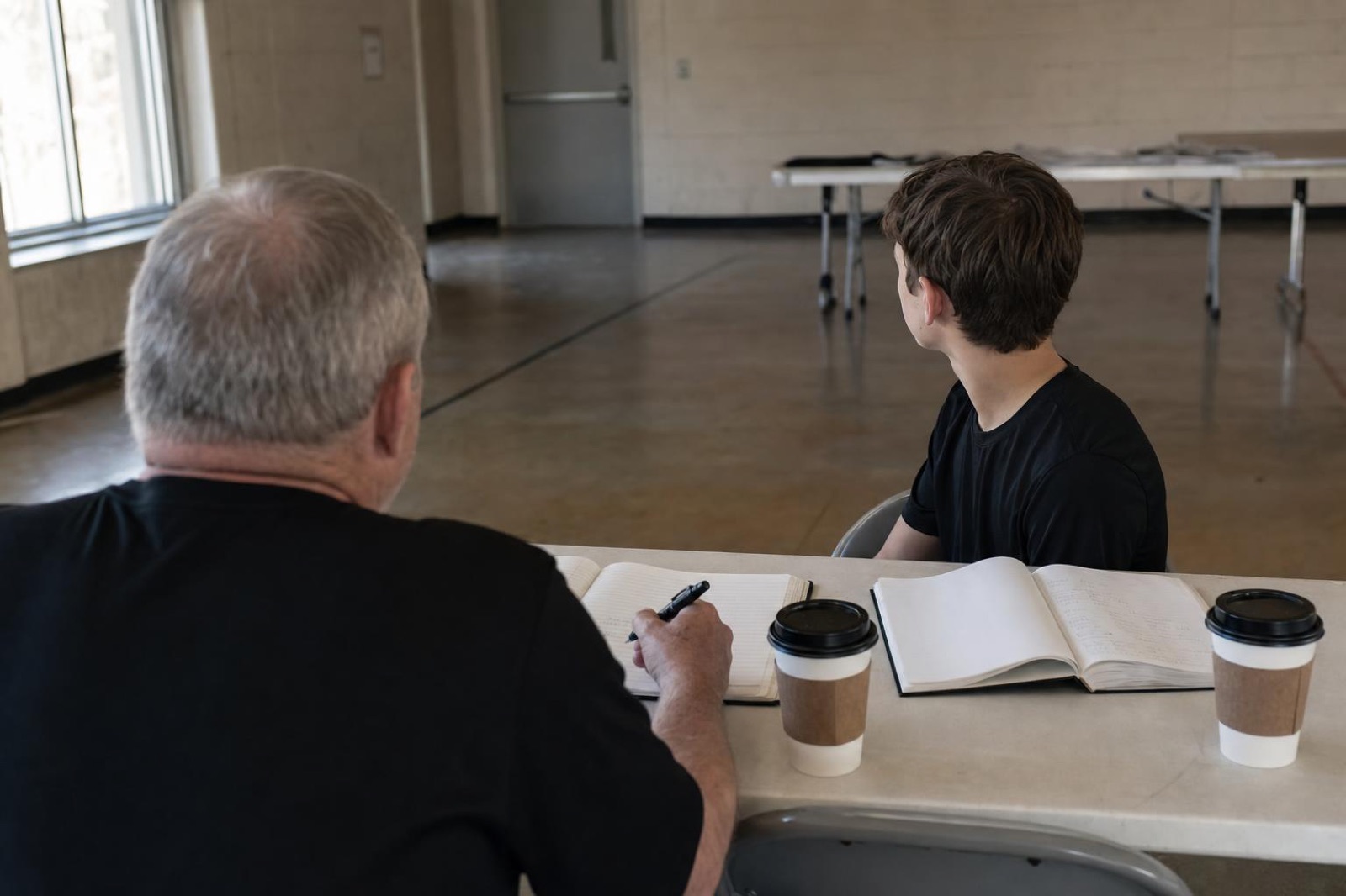 A Cadre member and a recruit talking across a folding table with notebooks and coffee, seen from behind