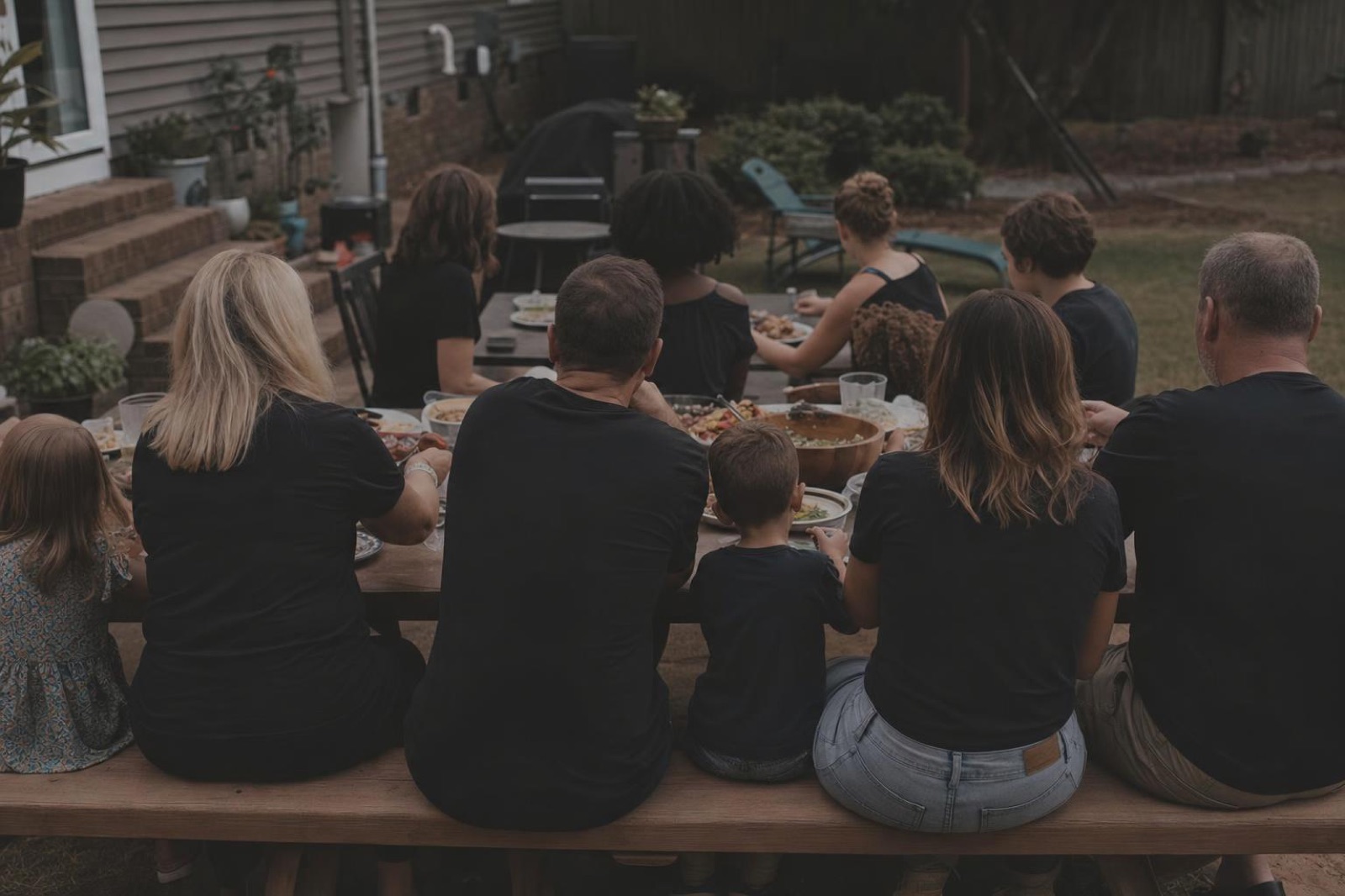 Two families sharing dinner at one long backyard table at dusk, seen from behind