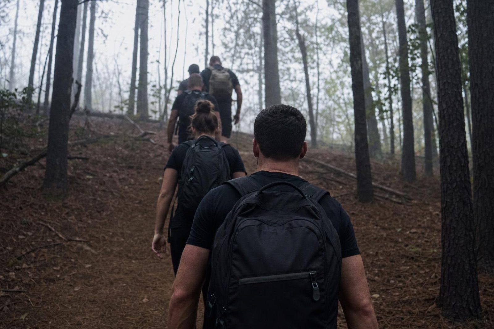 Single-file line of people with rucksacks climbing a misty forest trail, seen from behind