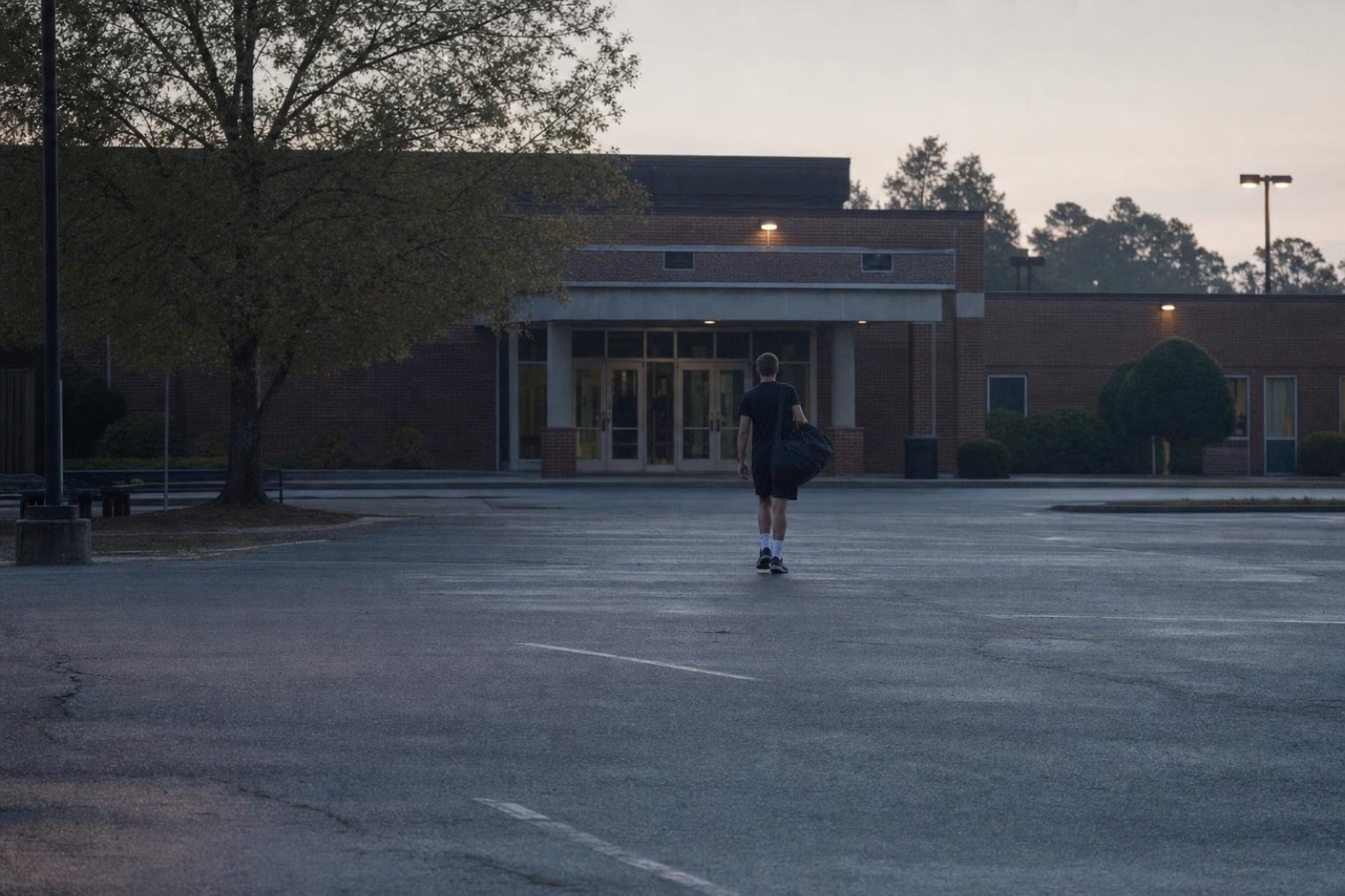 A young person with a duffel bag walking alone toward building doors at dawn, seen from behind